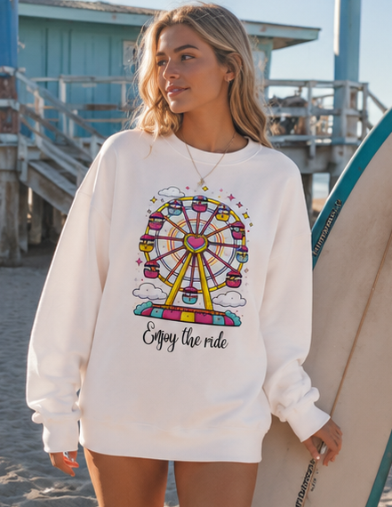 A woman on a beach wearing a white crewneck sweatshirt with a colorful Ferris wheel graphic and the text 'Enjoy the ride'. She is holding a surfboard next to her.