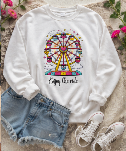 A white crewneck sweatshirt with a colorful Ferris wheel graphic and the text 'Enjoy the ride', styled in a flat lay with denim shorts, white sneakers, and pink flowers on a textured beige blanket.