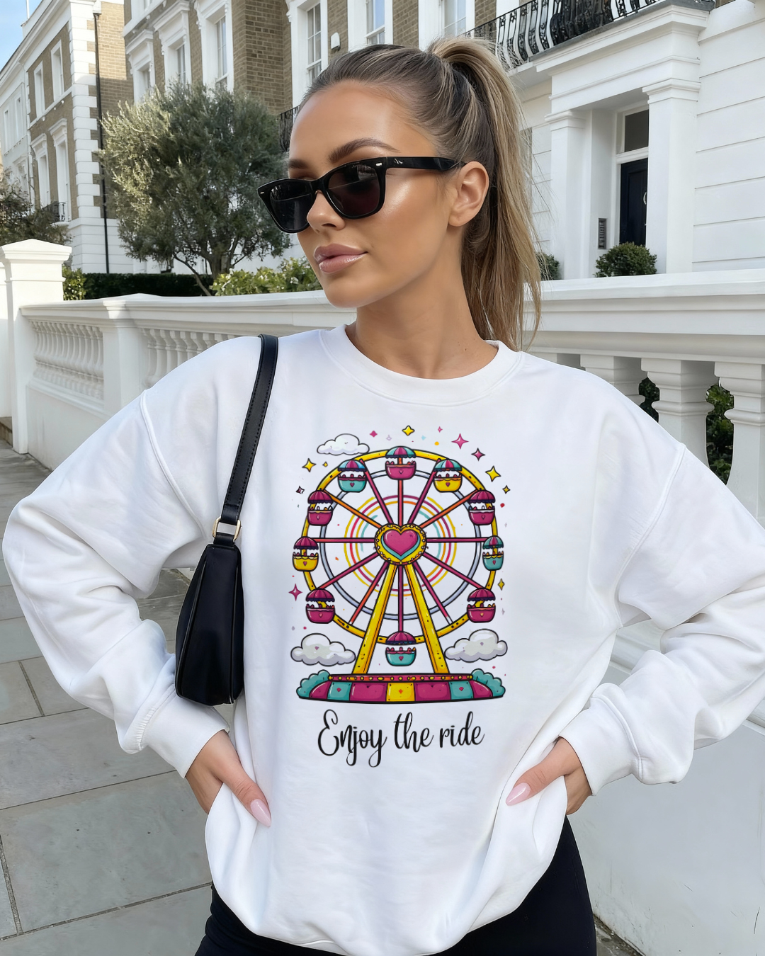 Woman wearing a white sweatshirt with a colorful ferris wheel design and 'Enjoy the ride' text, standing in an urban setting.