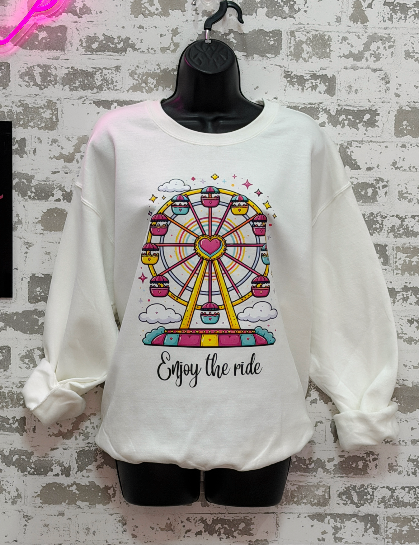 White sweatshirt with a colorful Ferris wheel graphic and 'Enjoy the ride' text on a textured wall background.