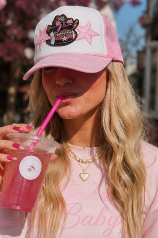 A woman wearing a pink and white trucker hat with a graphic patch, drinking from a pink straw cup outdoors.