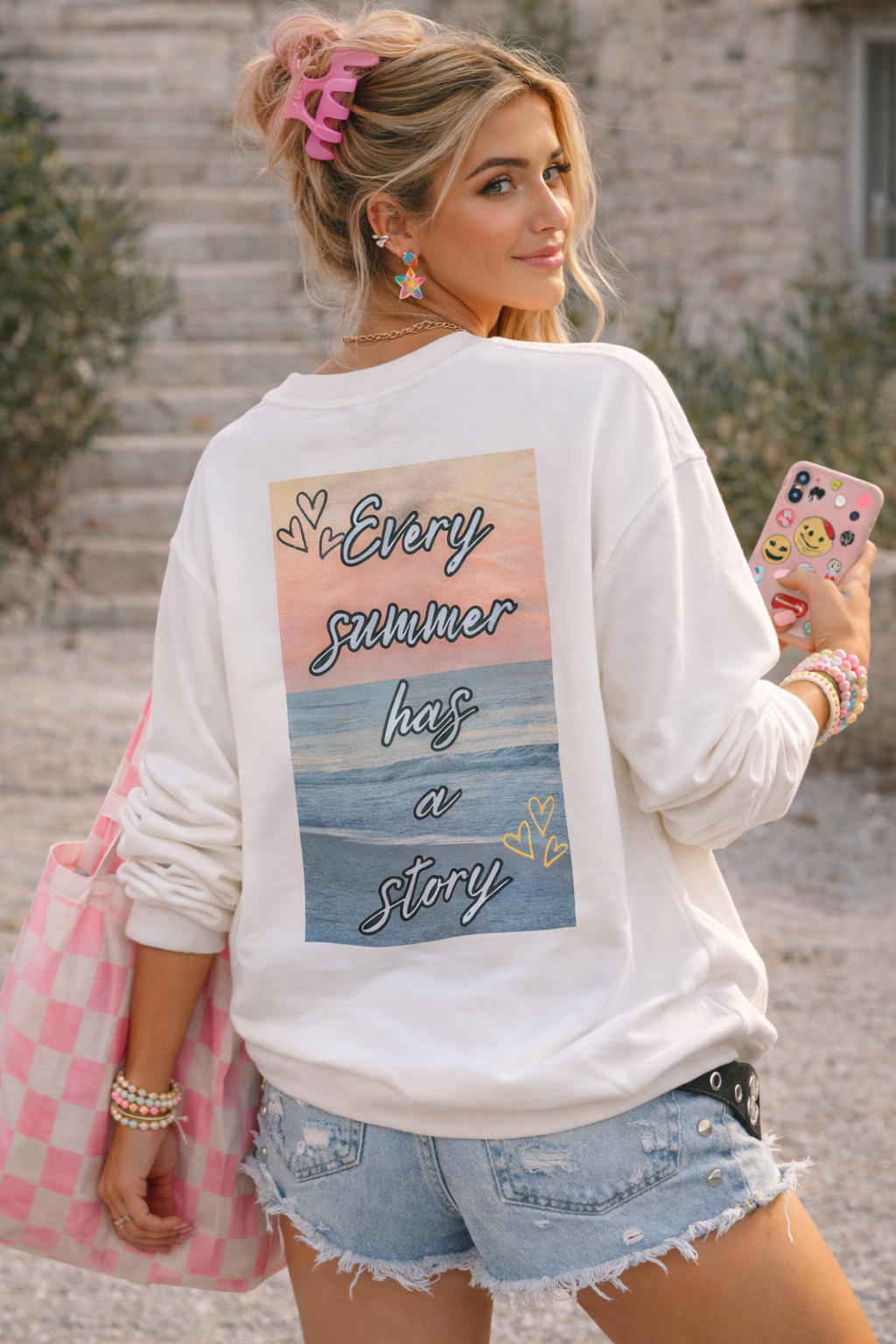 Woman wearing a white sweatshirt with a graphic design and text, holding a phone, standing outdoors.