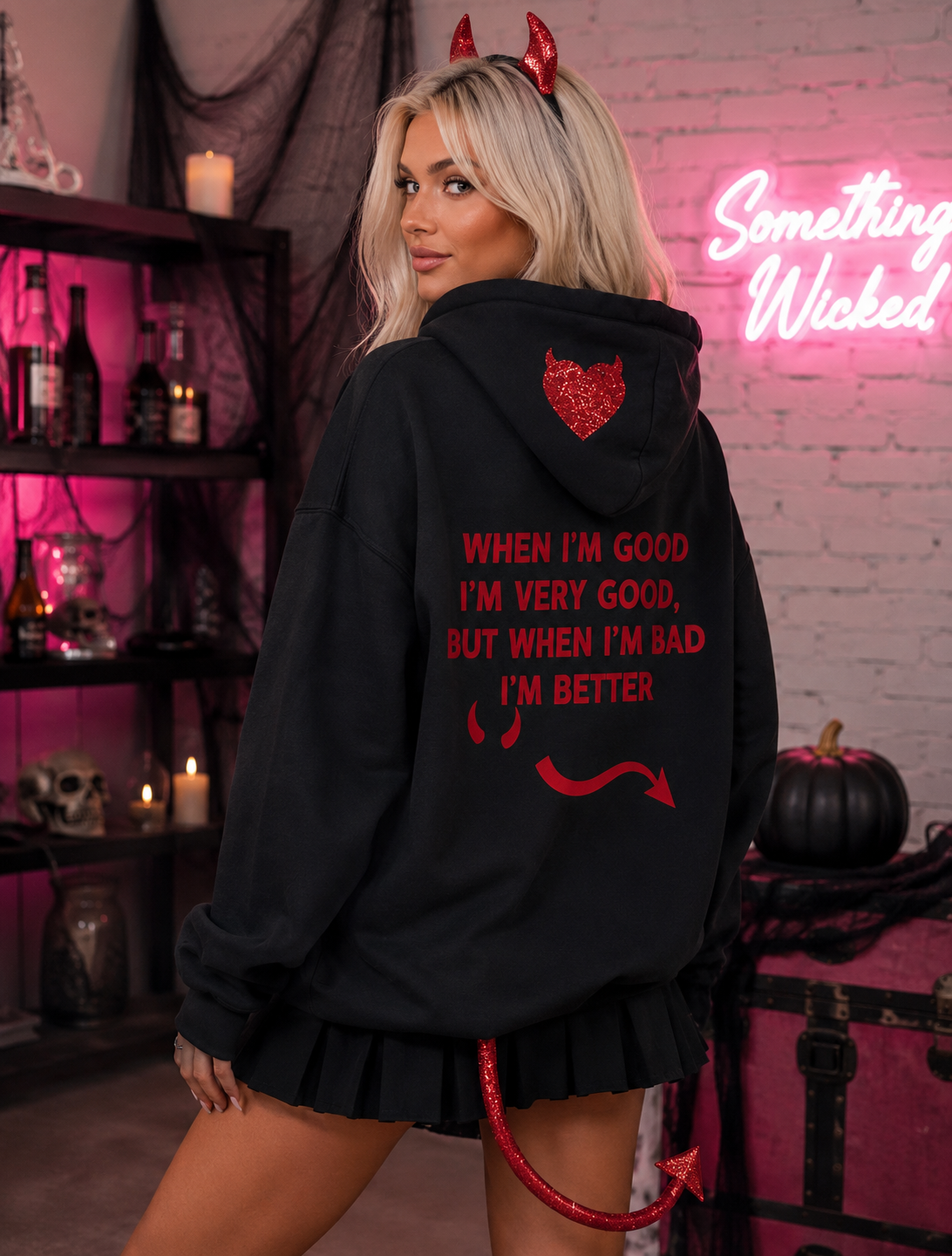 A woman wearing a black hoodie with red devil-themed text and graphics on the back, posed in a Halloween-themed room with a pink neon sign, candles, and spooky decor.
