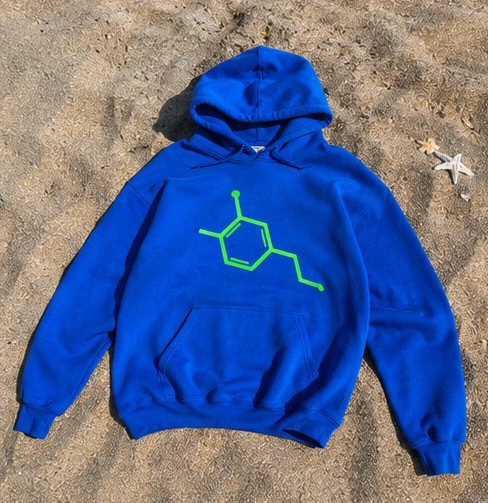 A bright blue pullover hoodie with a neon green chemical structure graphic on the chest, laid flat on a sandy beach with starfish nearby.