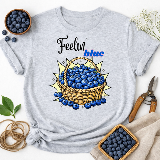 Gray t-shirt with a blueberry graphic and 'Feelin' blue' text, surrounded by blueberries and rustic items.