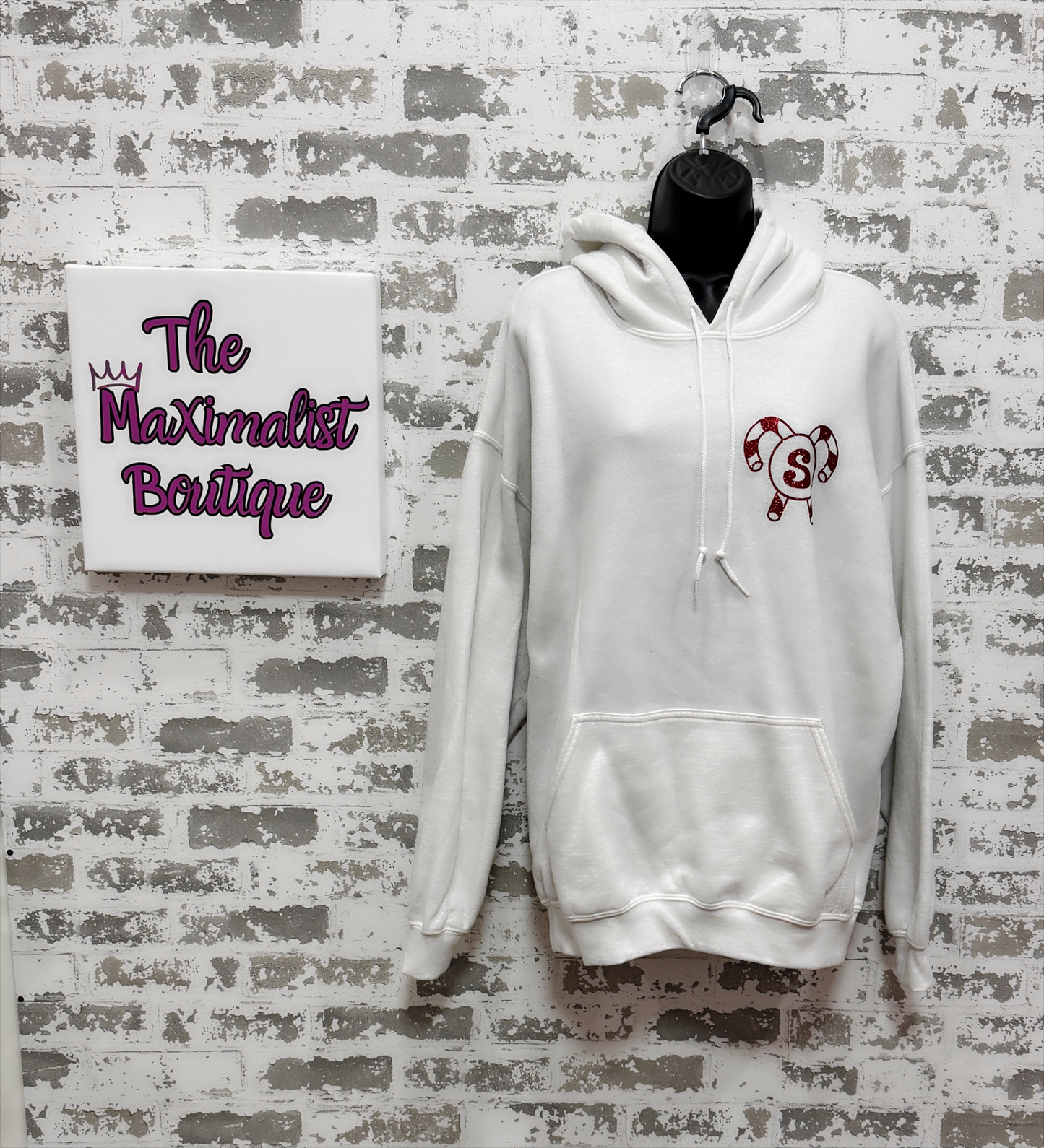 White hoodie with a logo on a gray brick wall with 'The Maximalist Boutique' sign.