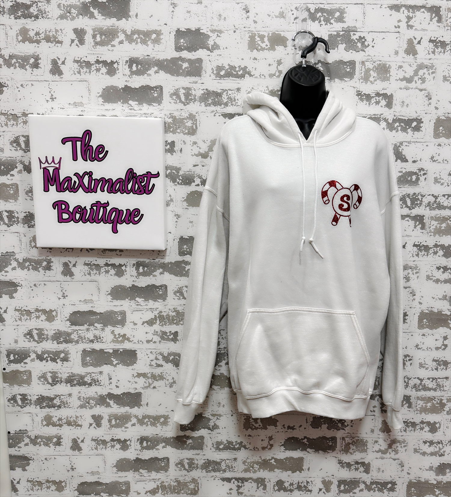 White hoodie with a logo on a gray brick wall with 'The Maximalist Boutique' sign.