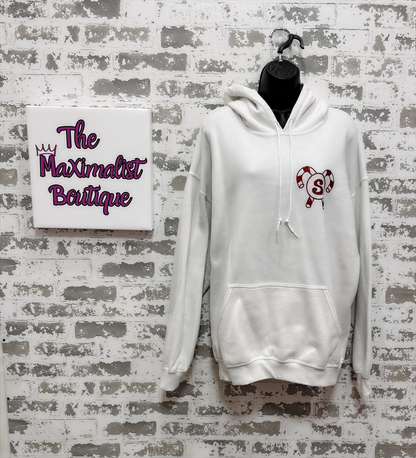 White hoodie with a logo on a gray brick wall with 'The Maximalist Boutique' sign.