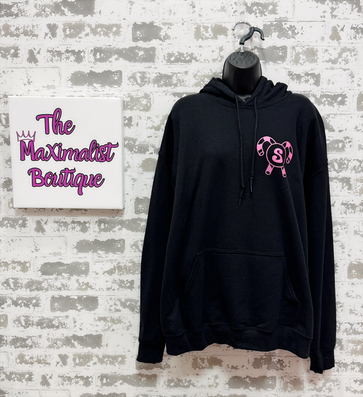 Black hoodie with pink logo on a mannequin against a textured wall with 'The Maximalist Boutique' sign.