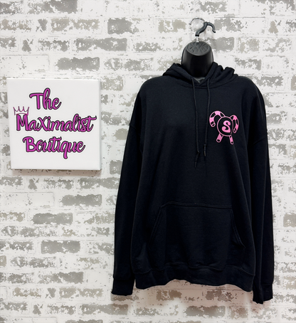 Black hoodie with pink logo on a mannequin against a textured wall with 'The Maximalist Boutique' sign.