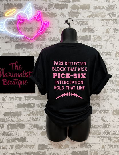 A black short-sleeve T-shirt displayed on a black mannequin torso against a faux white brick wall. The back of the shirt features pink text reading 'PASS DEFLECTED BLOCK THAT KICK PICK-SIX INTERCEPTION HOLD THAT LINE' with a pink football graphic below.