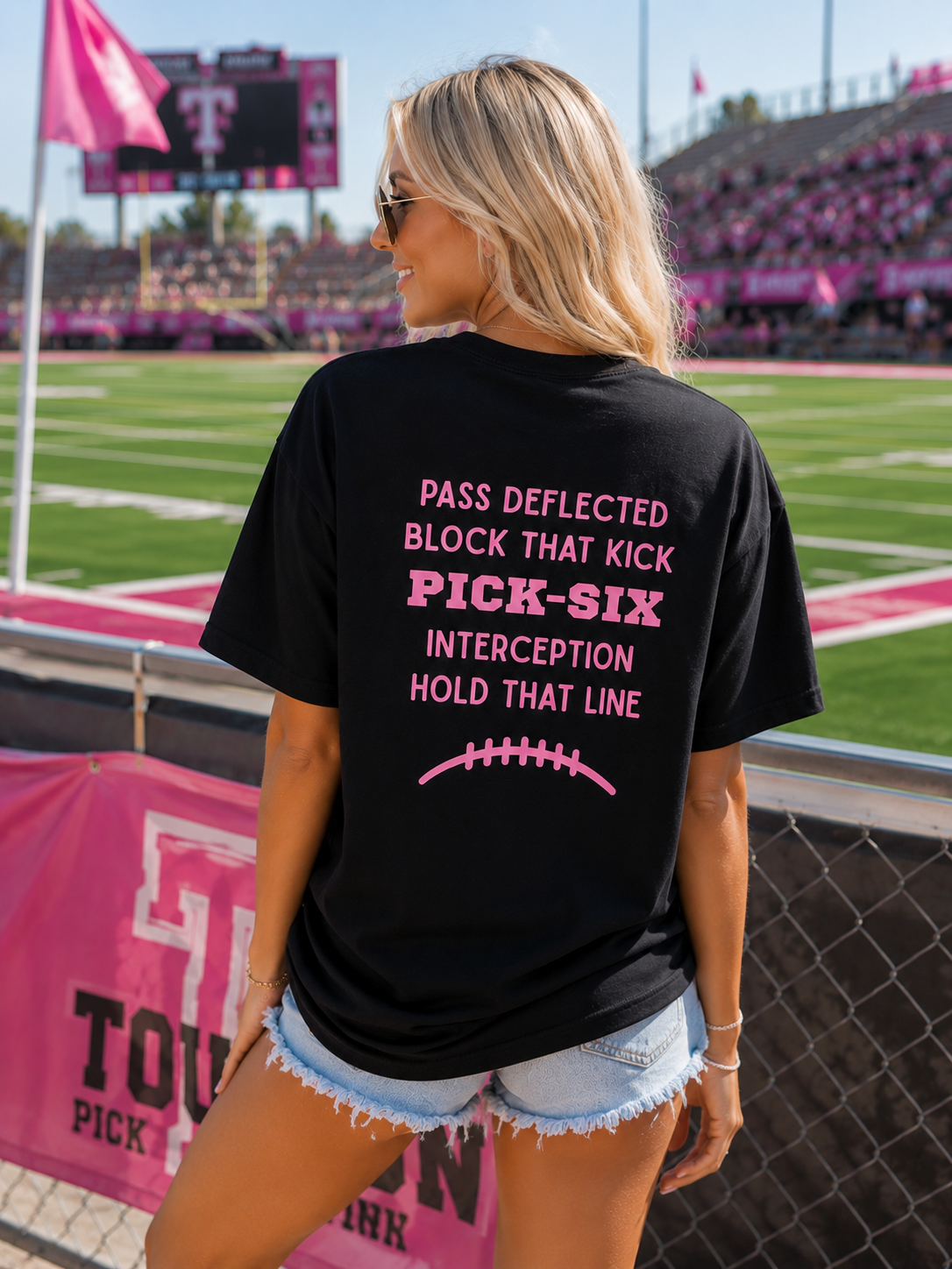A woman standing on a football field wearing a black graphic t-shirt with pink text that reads 'PASS DEFLECTED BLOCK THAT KICK PICK-SIX INTERCEPTION HOLD THAT LINE' and a pink football graphic.