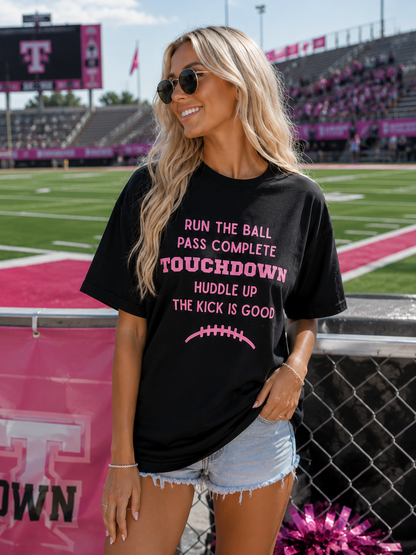 A woman wearing a black graphic t-shirt with pink text that reads 'RUN THE BALL PASS COMPLETE TOUCHDOWN HUDDLE UP THE KICK IS GOOD' and a pink football graphic, standing on a football field sideline.