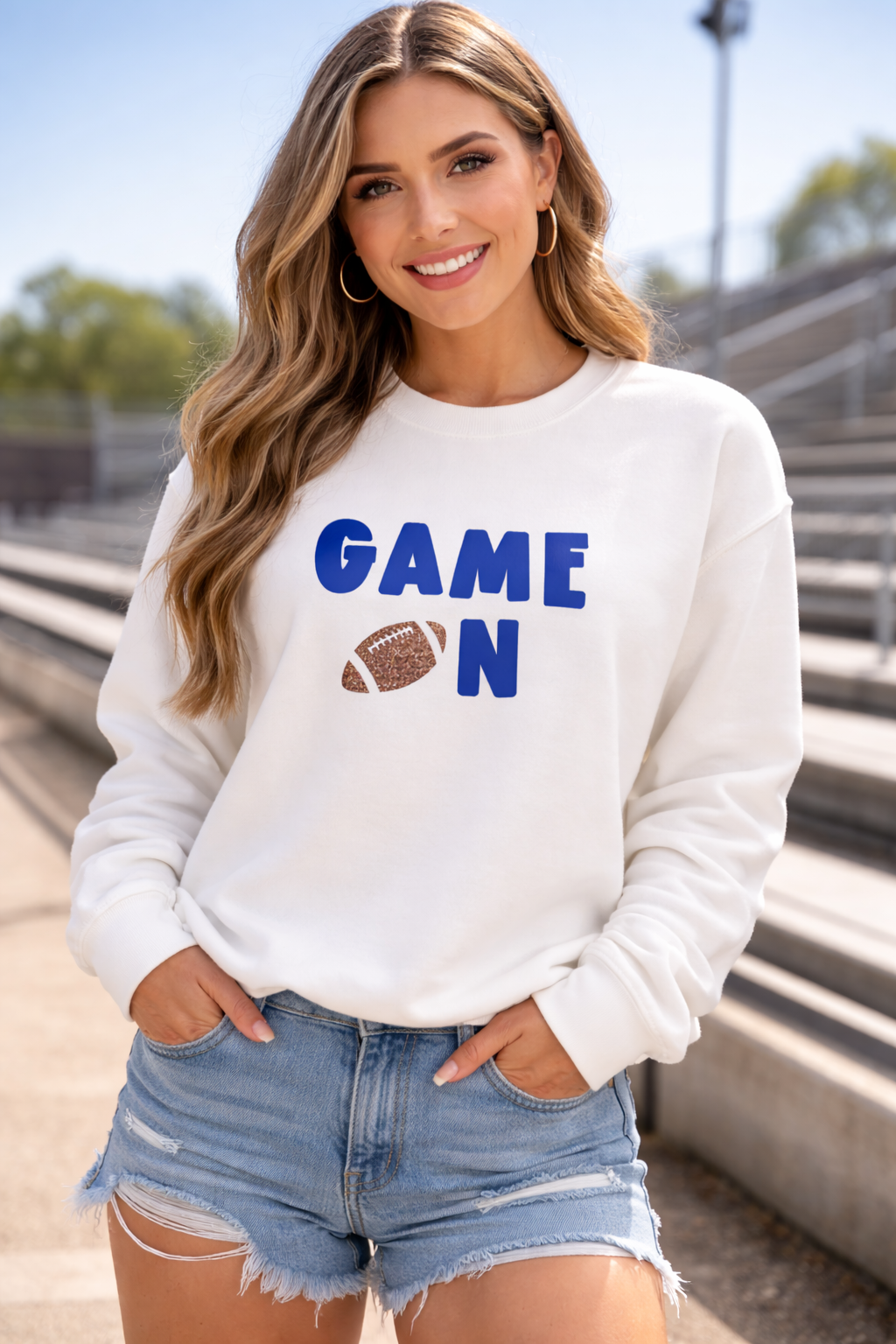 Woman wearing a white sweatshirt with 'GAME ON' and a football graphic, standing outdoors.