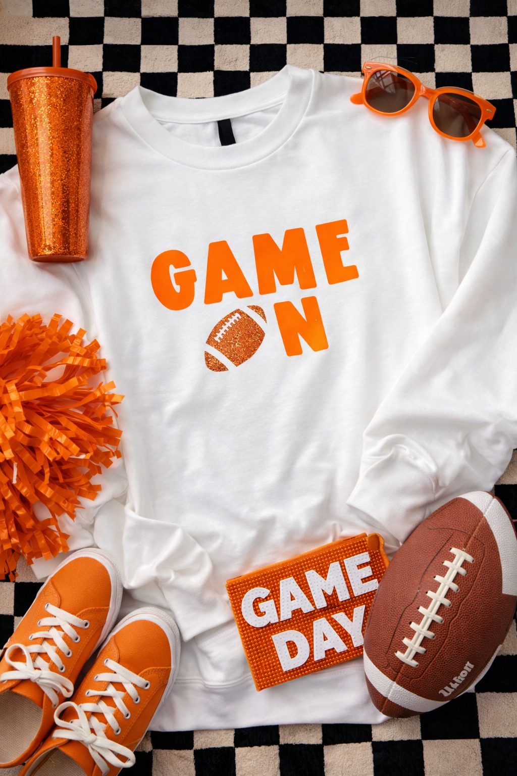 White sweatshirt with 'GAME ON' text, orange sneakers, football, pom-pom, and sunglasses on a checkered background.
