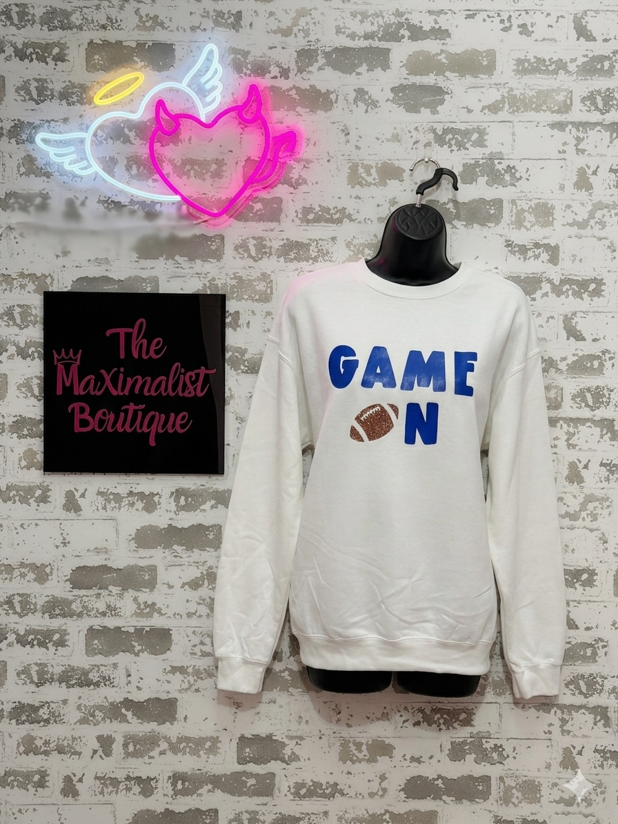 White sweatshirt with 'GAME ON' text, neon sign, and 'The Maximalist Boutique' sign on a textured wall.