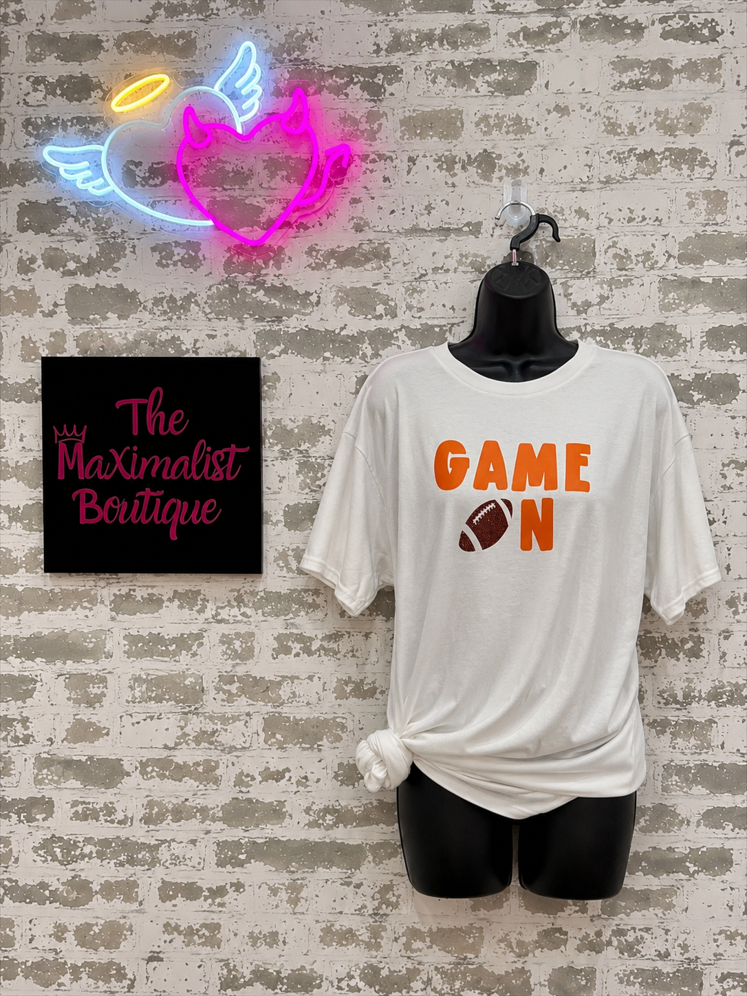 Neon sign, t-shirt with 'GAME ON' and football graphic, and 'The Maximalist Boutique' sign on a textured wall.