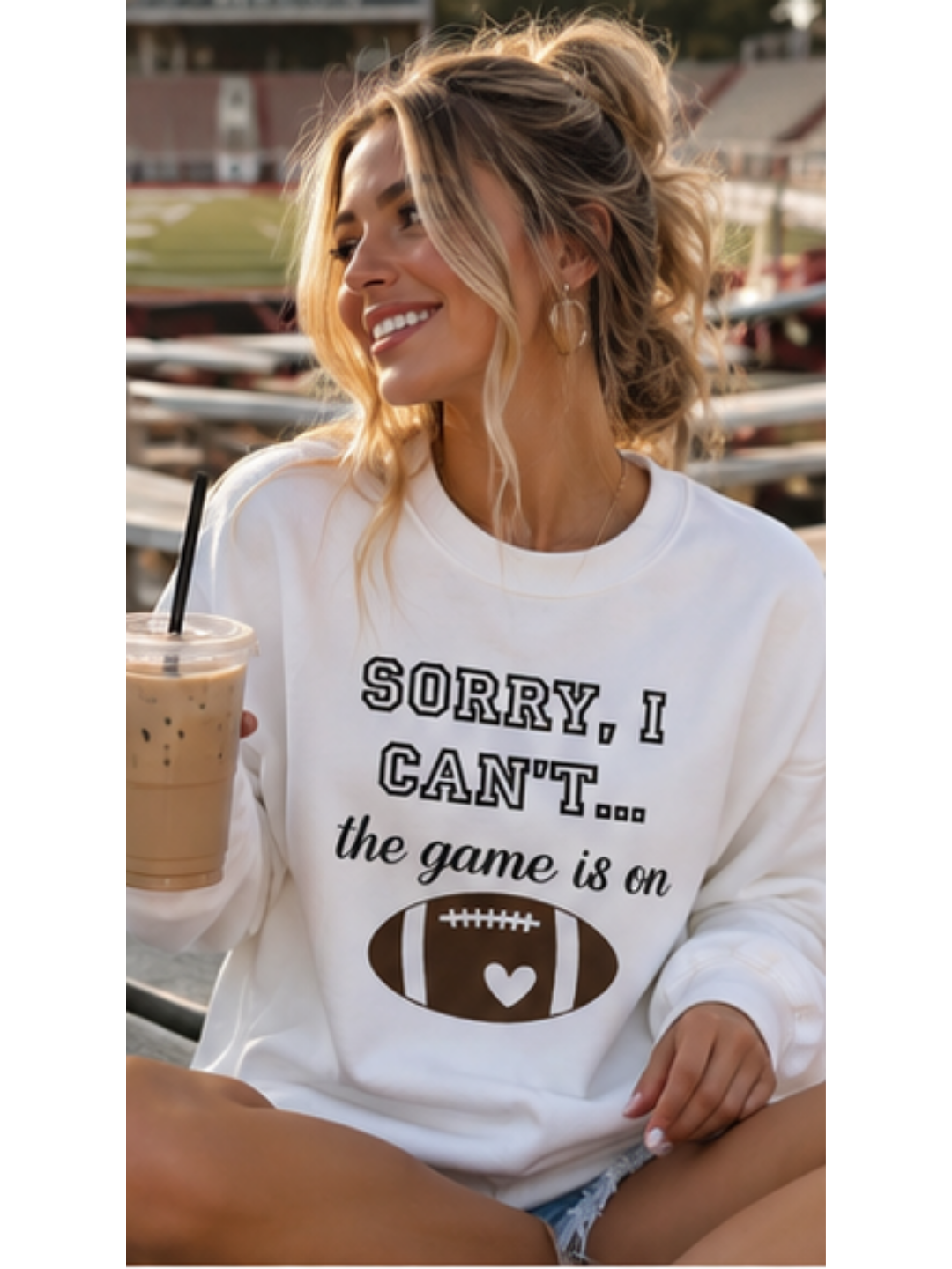 Woman wearing a sweatshirt with a football graphic and text, holding a coffee in a stadium.
