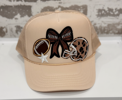 Beige cap with decorative patches including a bow, football, helmet, and star on a white surface.