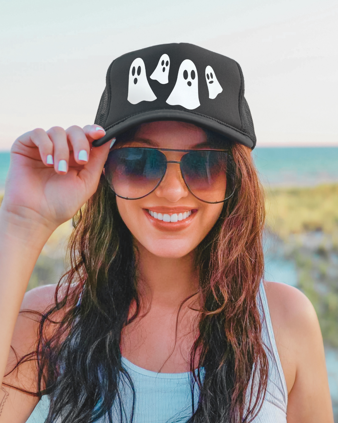 Woman wearing a black cap with ghost designs and sunglasses, standing by the beach.