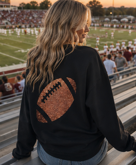 A woman sitting in stadium bleachers at sunset, viewed from behind, wearing a black sweatshirt with a large glittery copper football graphic on the back.