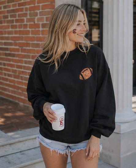 A woman wearing a black crewneck sweatshirt with a brown football graphic on the chest, holding a white coffee cup, standing outdoors in front of a brick wall and white columns.