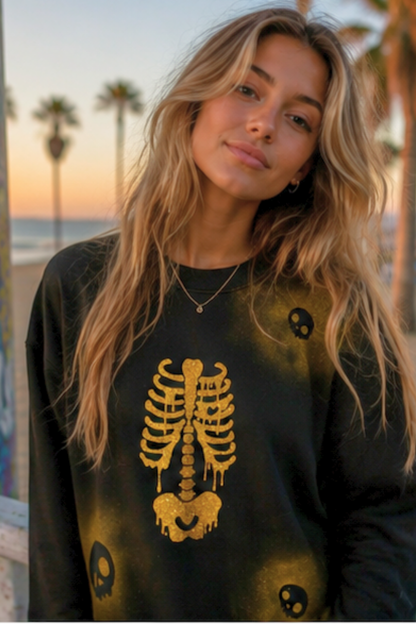 A woman with long blonde hair wearing a black crewneck sweatshirt with a gold dripping skeleton graphic and skull prints, photographed outdoors against a sunset beach background with palm trees.