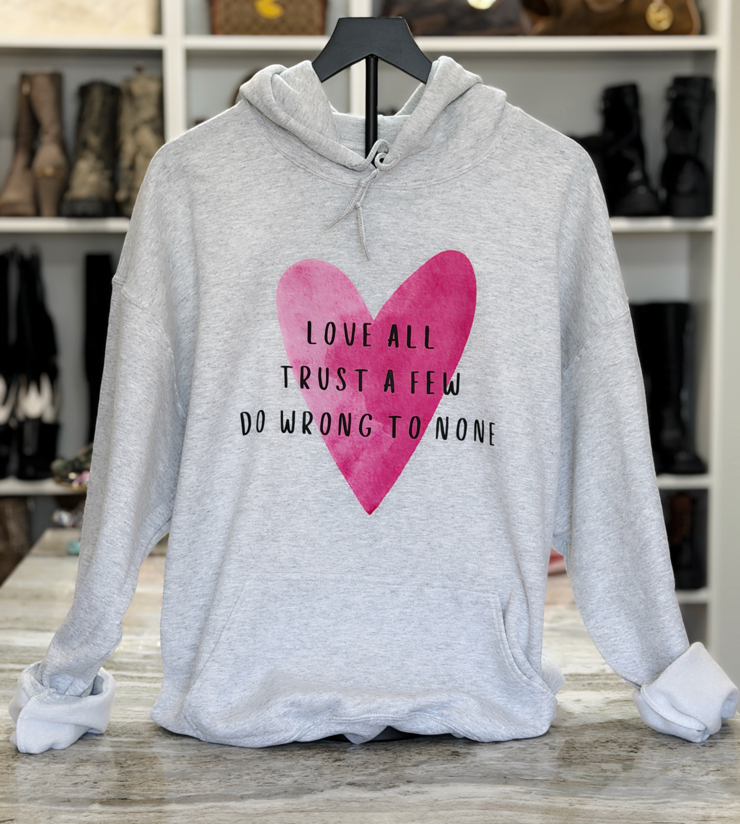 Gray hoodie with pink heart and text on a wooden surface with shelves in the background