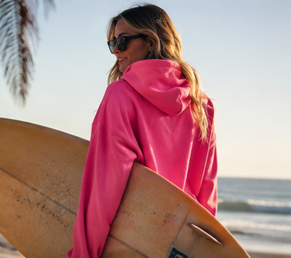 A woman on a beach wearing a bright pink hooded sweatshirt and sunglasses, holding a surfboard.