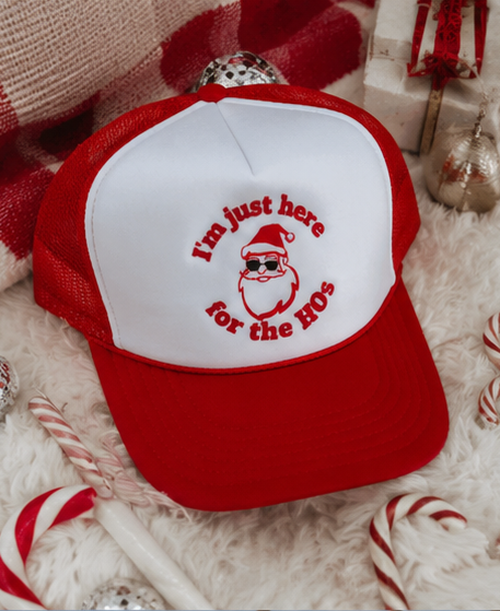 A red and white trucker hat with a Santa graphic and the text 'I'm just here for the HOs' on the front panel, styled on a white faux fur surface with candy canes and holiday decorations.