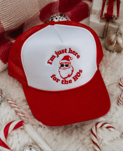 A red and white trucker hat with a Santa graphic and the text 'I'm just here for the HOs' on the front panel, styled on a white faux fur surface with candy canes and holiday decorations.