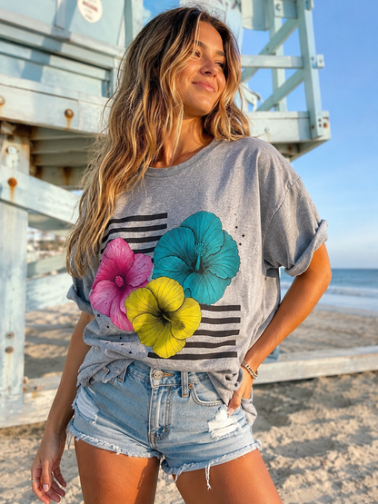 A woman standing on a beach in front of a wooden lifeguard tower, wearing a heather gray graphic t-shirt with a black stripe pattern and three large hibiscus flowers in pink, teal, and yellow, paired with light-wash denim shorts.