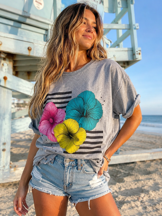 A woman standing on a beach in front of a wooden lifeguard tower, wearing a heather gray graphic t-shirt with a black stripe pattern and three large hibiscus flowers in pink, teal, and yellow, paired with light-wash denim shorts.