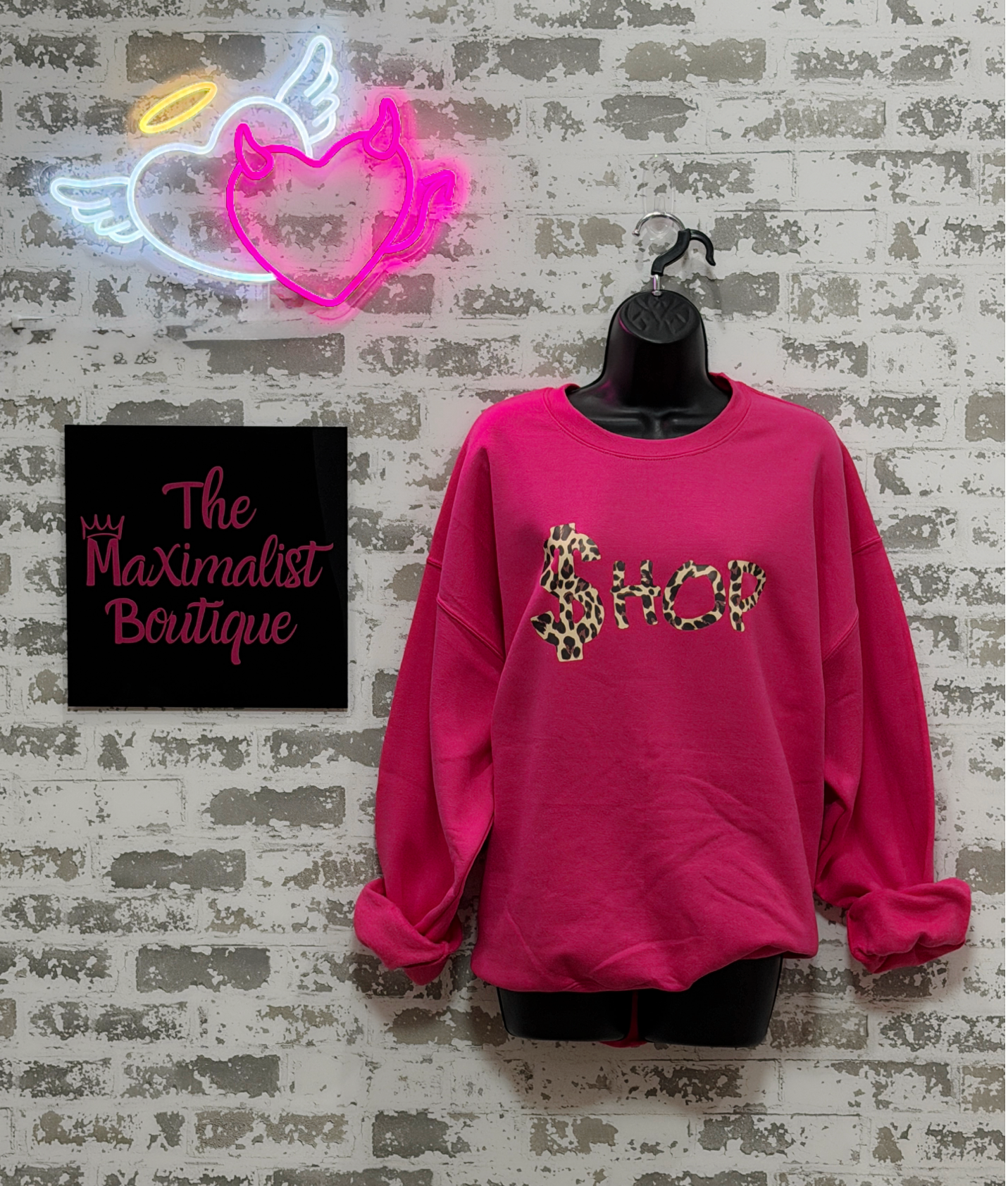 Pink sweatshirt with leopard print text on a mannequin against a textured wall with neon sign and 'The Maximalist Boutique' sign.