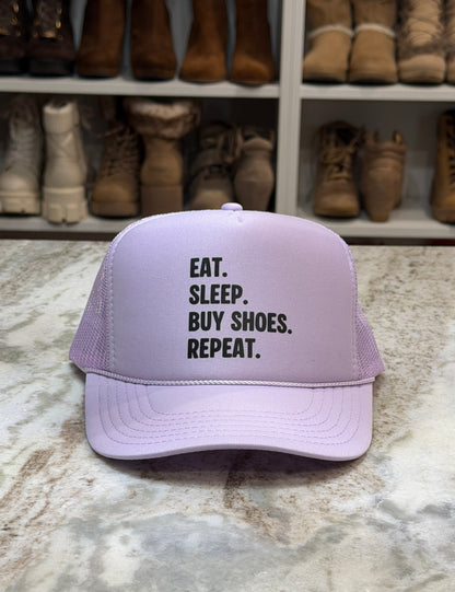 Lavender cap with 'Eat. Sleep. Buy Shoes. Repeat.' text on a marble surface with shoes in the background.
