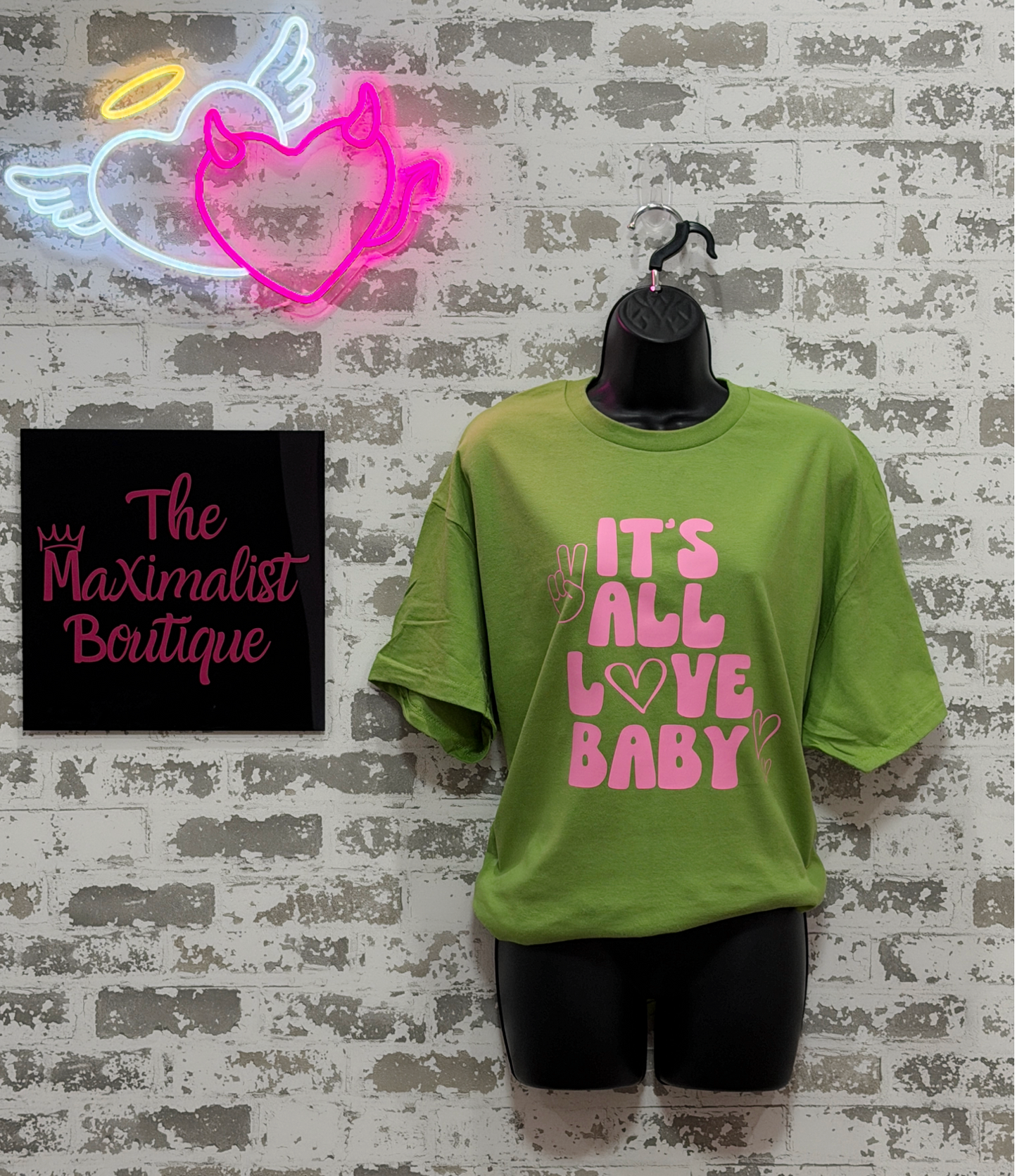 Green t-shirt with pink text 'IT'S ALL LOVE BABY' on a mannequin against a textured wall with neon sign and 'The Maximalist Boutique' sign.