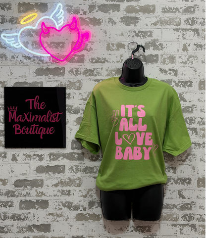 Green t-shirt with pink text 'IT'S ALL LOVE BABY' on a mannequin against a textured wall with neon sign and 'The Maximalist Boutique' sign.