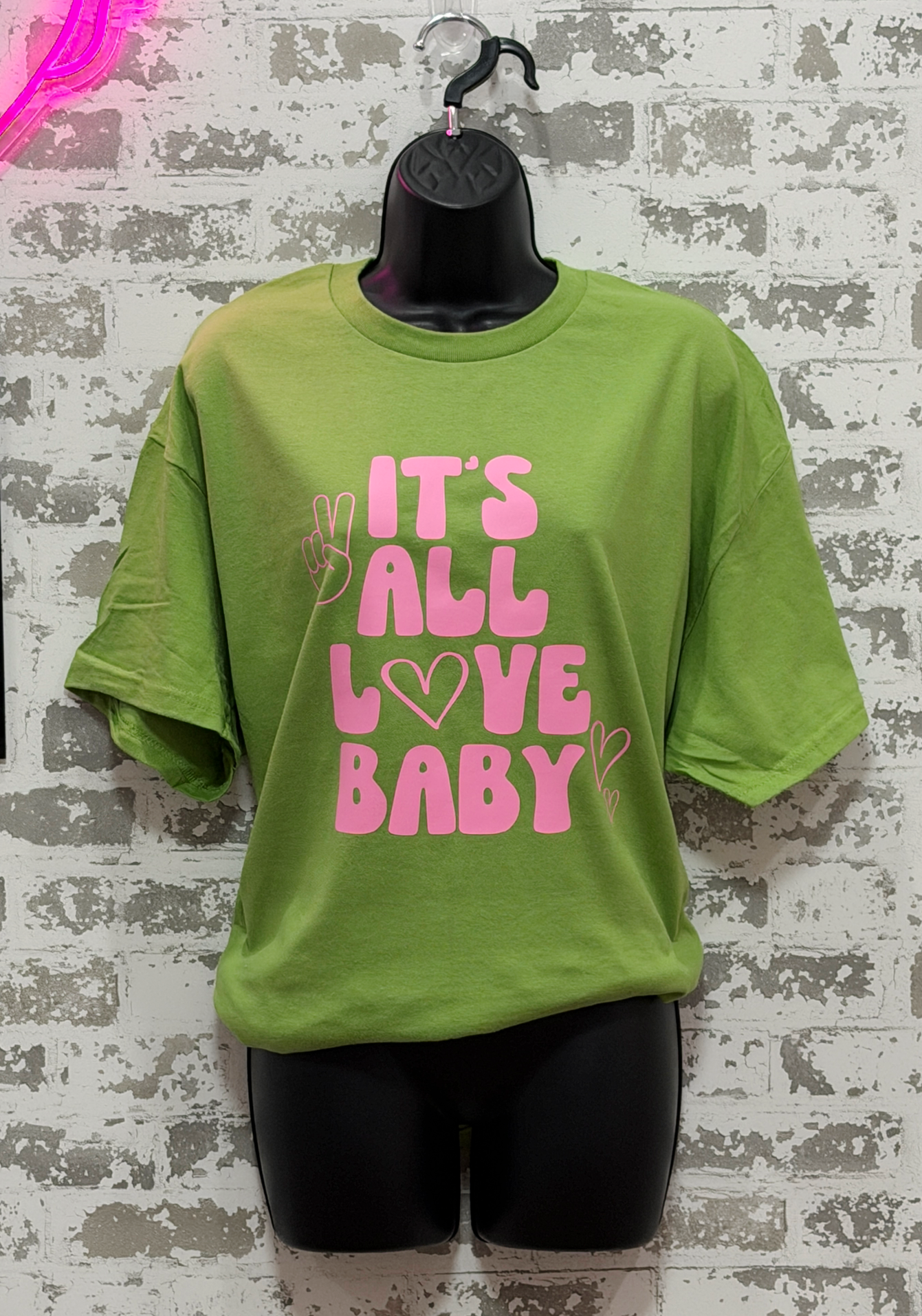 Green t-shirt with pink text 'IT'S ALL LOVE BABY' on a textured wall background