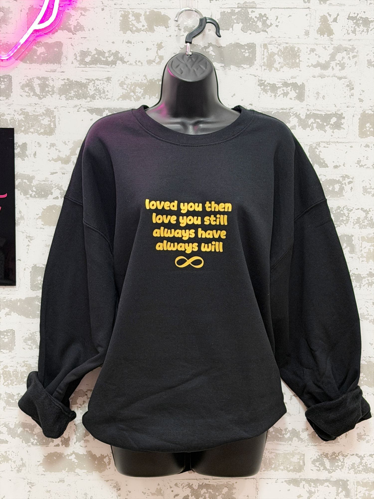 Black sweatshirt with gold text on a hanger against a textured wall.