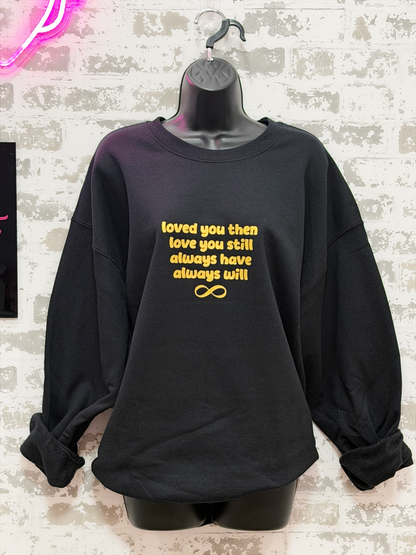Black sweatshirt with gold text on a hanger against a textured wall.