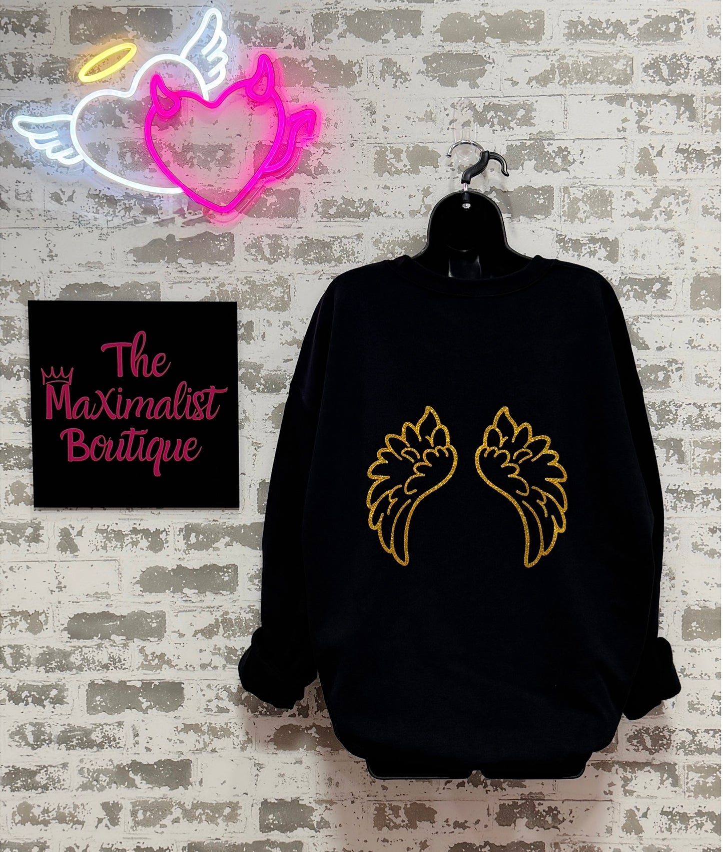 Black sweatshirt with gold angel wings design on a hanger against a textured wall with 'The Maximalist Boutique' sign.