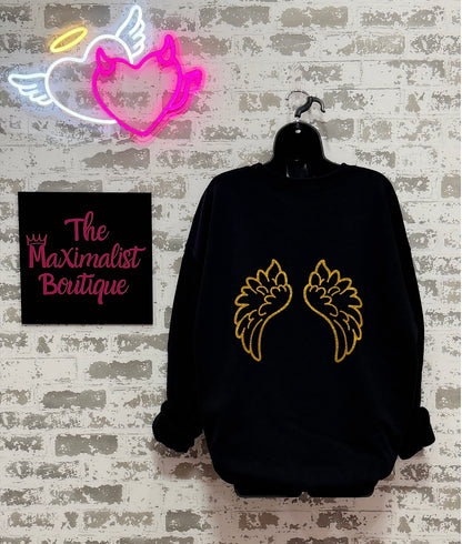 Black sweatshirt with gold angel wings design on a hanger against a textured wall with 'The Maximalist Boutique' sign.