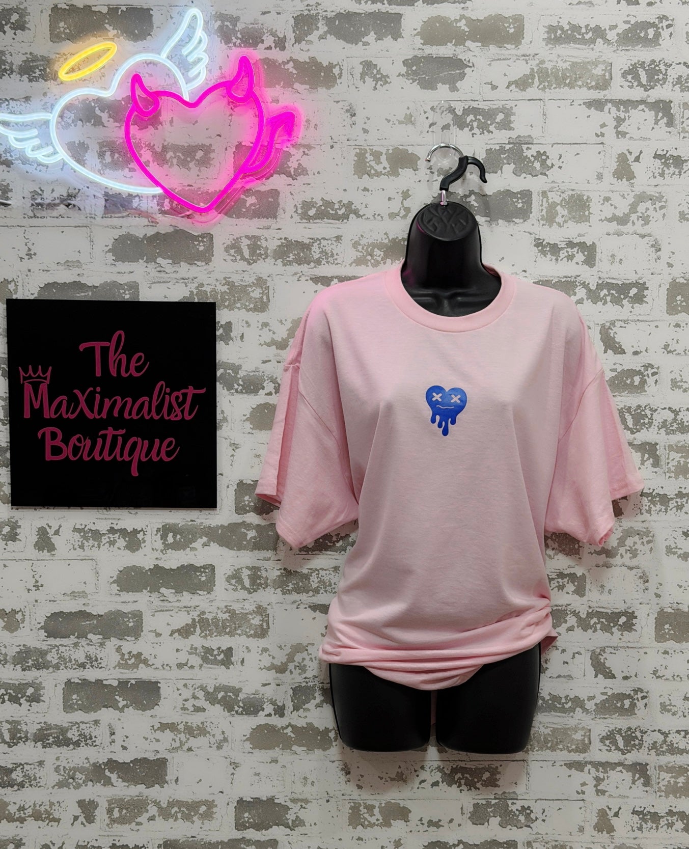 Pink t-shirt with a blue heart design on a mannequin against a textured wall with neon signs and 'The Maximalist Boutique' sign.