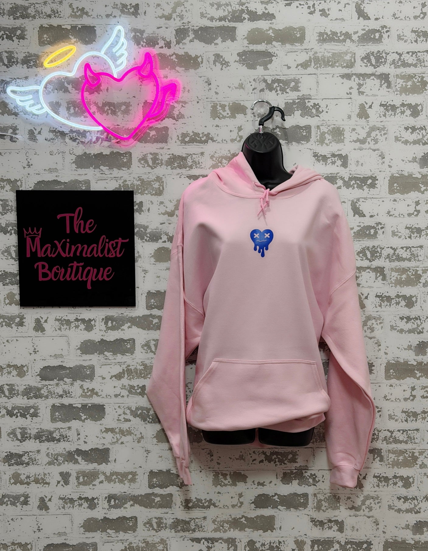 Pink hoodie on a mannequin against a textured wall with 'The Maximalist Boutique' sign.