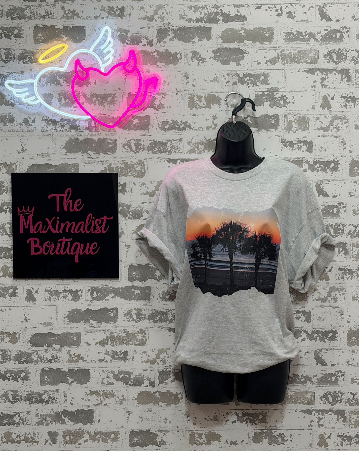 Gray t-shirt with palm tree design on a mannequin against a brick wall with a neon sign and 'The Maximalist Boutique' sign.