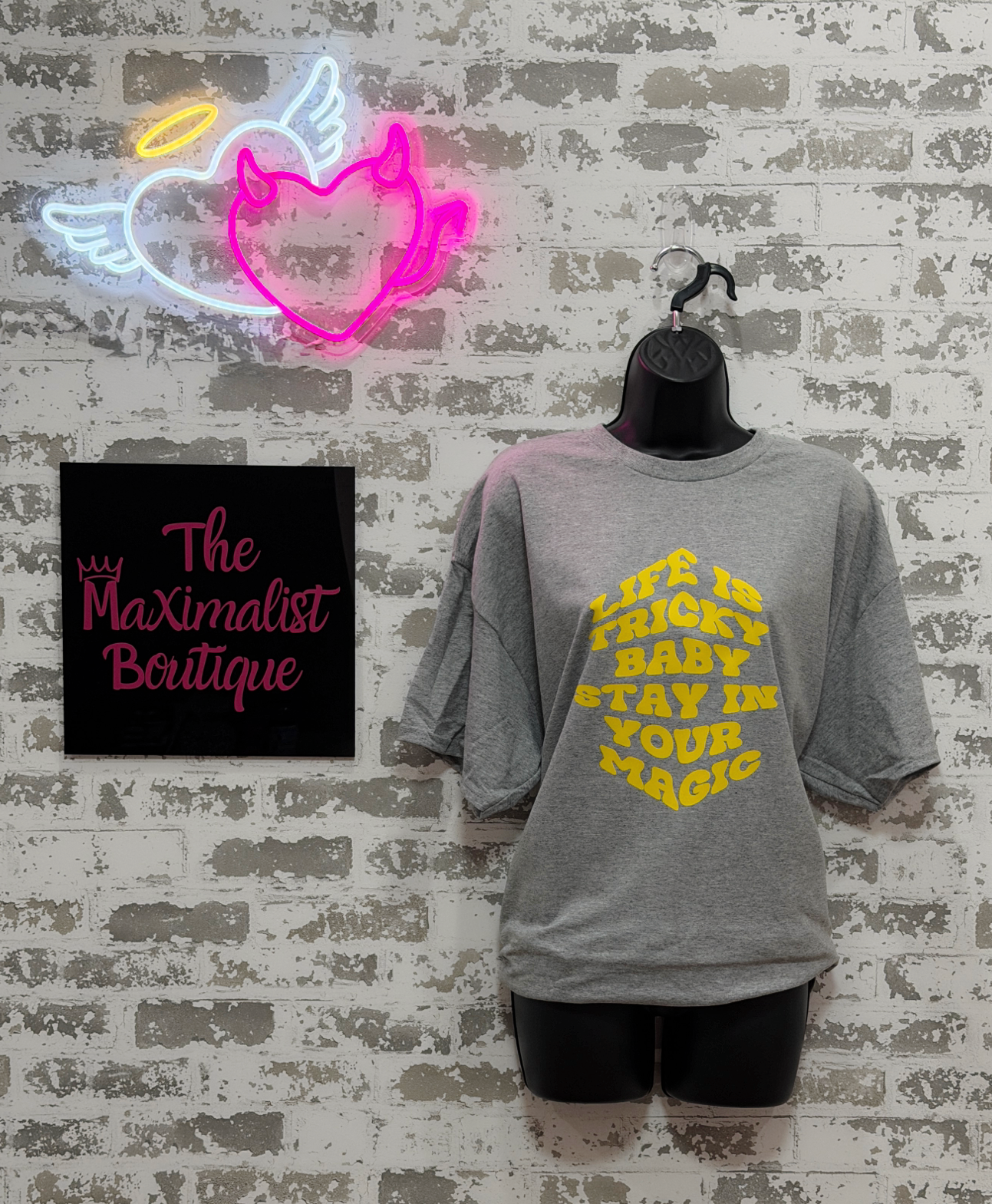 Gray t-shirt with yellow text on a mannequin against a textured wall with neon sign and 'The Maximalist Boutique' sign.