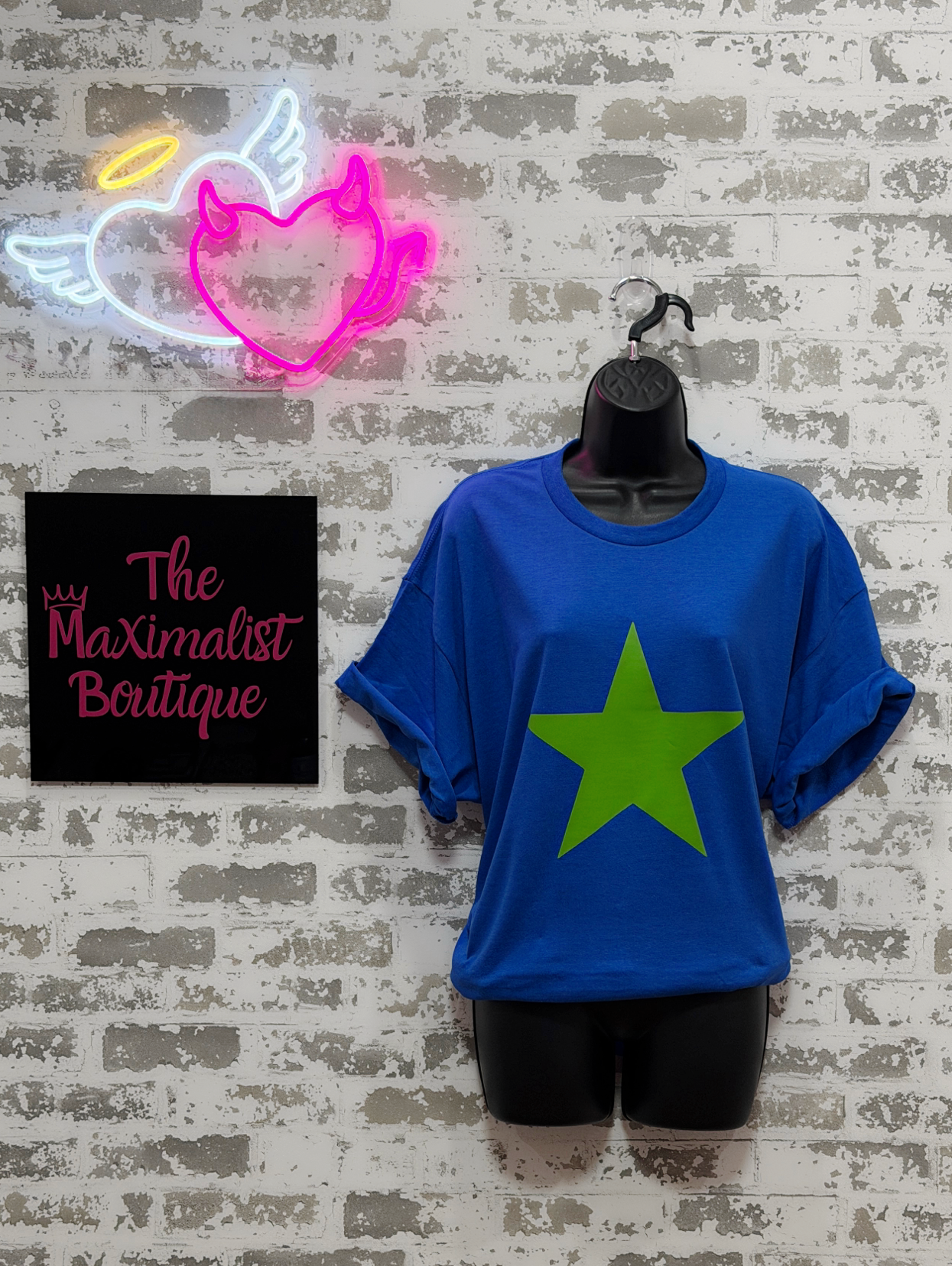 Blue t-shirt with a green star on a hanger against a textured wall with neon signs.