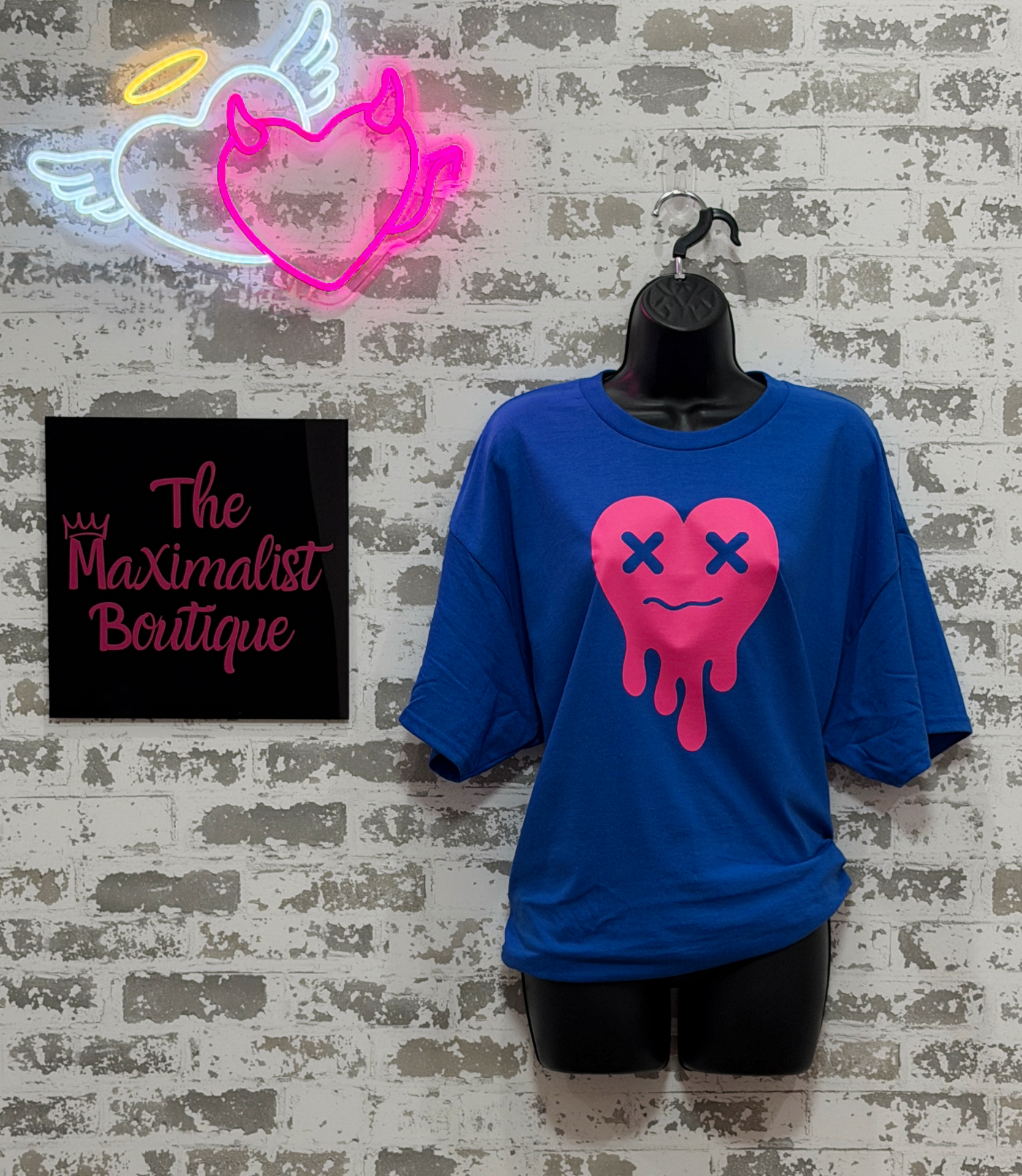 Blue t-shirt with pink heart design on a mannequin against a textured wall with neon sign and 'The Maximalist Boutique' sign.