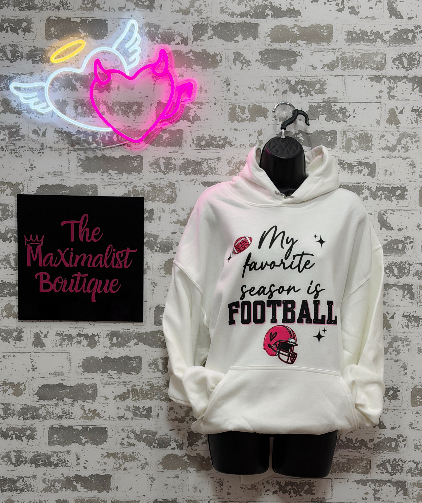 A white pullover hoodie displayed on a black mannequin torso against a distressed gray brick wall. The hoodie features a graphic print that reads 'My favorite season is FOOTBALL' in black and dark red lettering, accompanied by a football and helmet illustration.