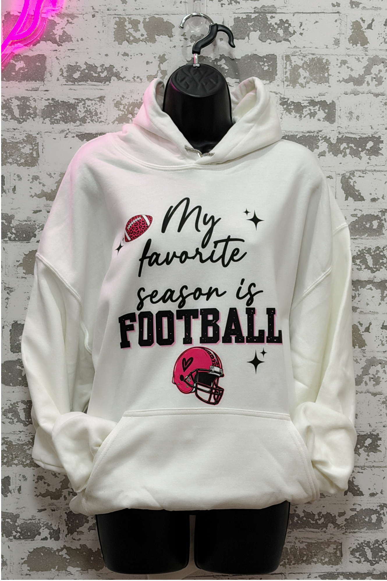 A white pullover hoodie displayed on a black mannequin torso against a distressed gray brick wall. The hoodie features a graphic print that reads 'My favorite season is FOOTBALL' in black script and block lettering, accompanied by illustrations of a red football and a red football helmet.